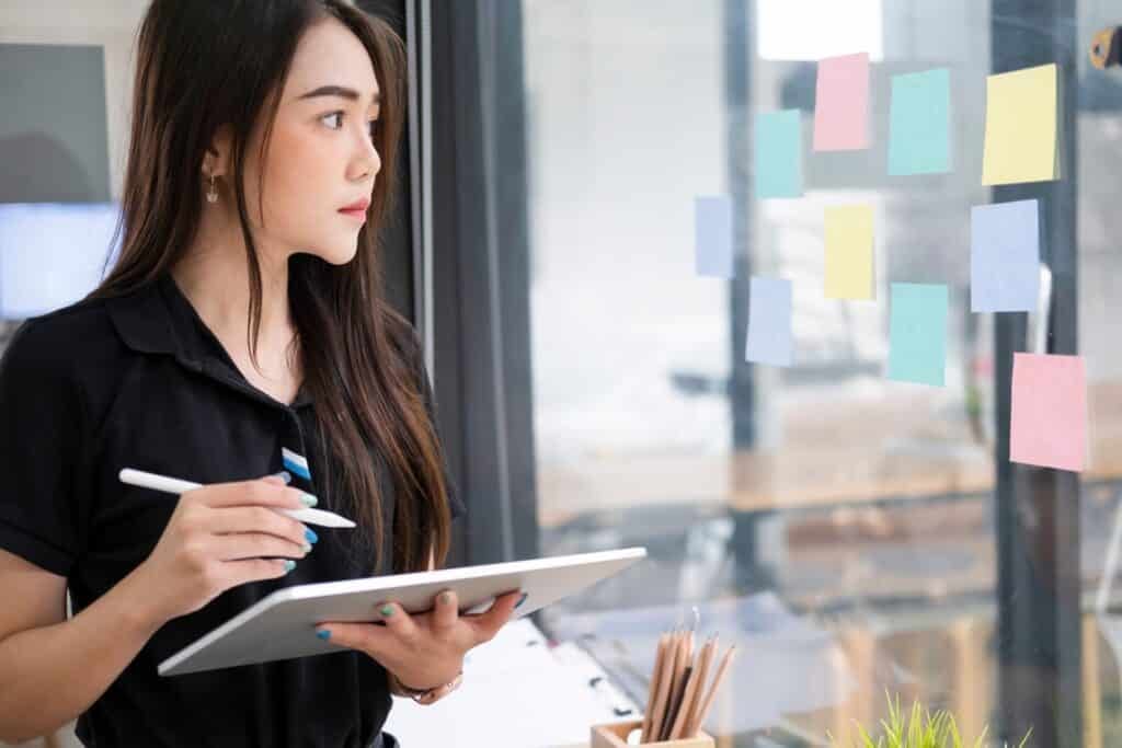 A woman taking notes on a tablet and using sticky notes on a window she is looking out. 