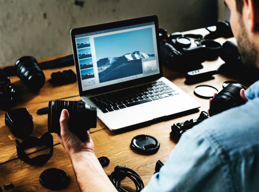 A man editing photos on a computer and holding a camera lens. 