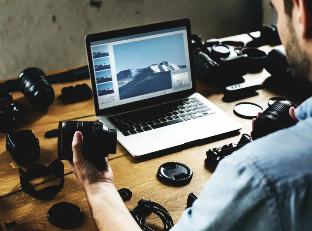 A man editing photos on a computer and holding a camera lens. 
