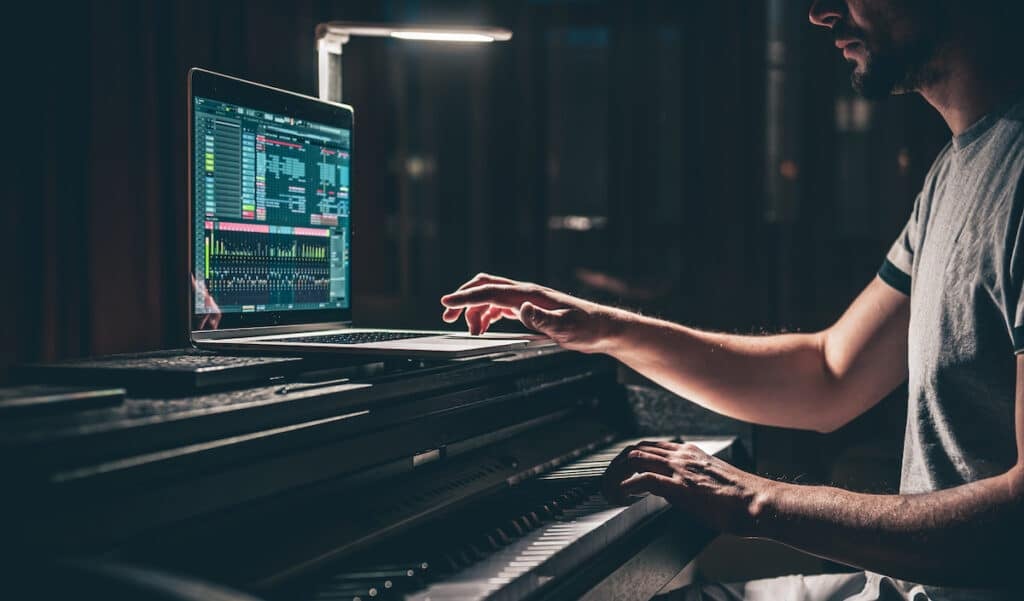 A man using a laptop sitting on a piano to produce music.